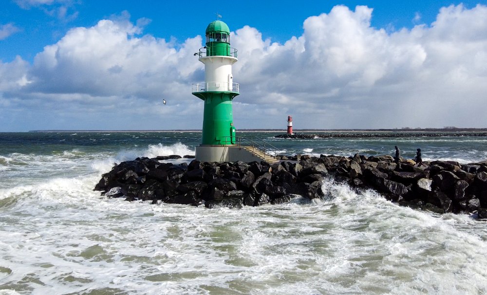 Das grün-weiße Molenfeuer der Warnemünder Westmole bei stürmischer Ostsee, Wellen mit Gischt schlagen gegen den Molenkopf, darüber ein aufgelockerter blauer Himmel mit weißen Wolken, im Hintergrund das rote Ostmolenfeuer.